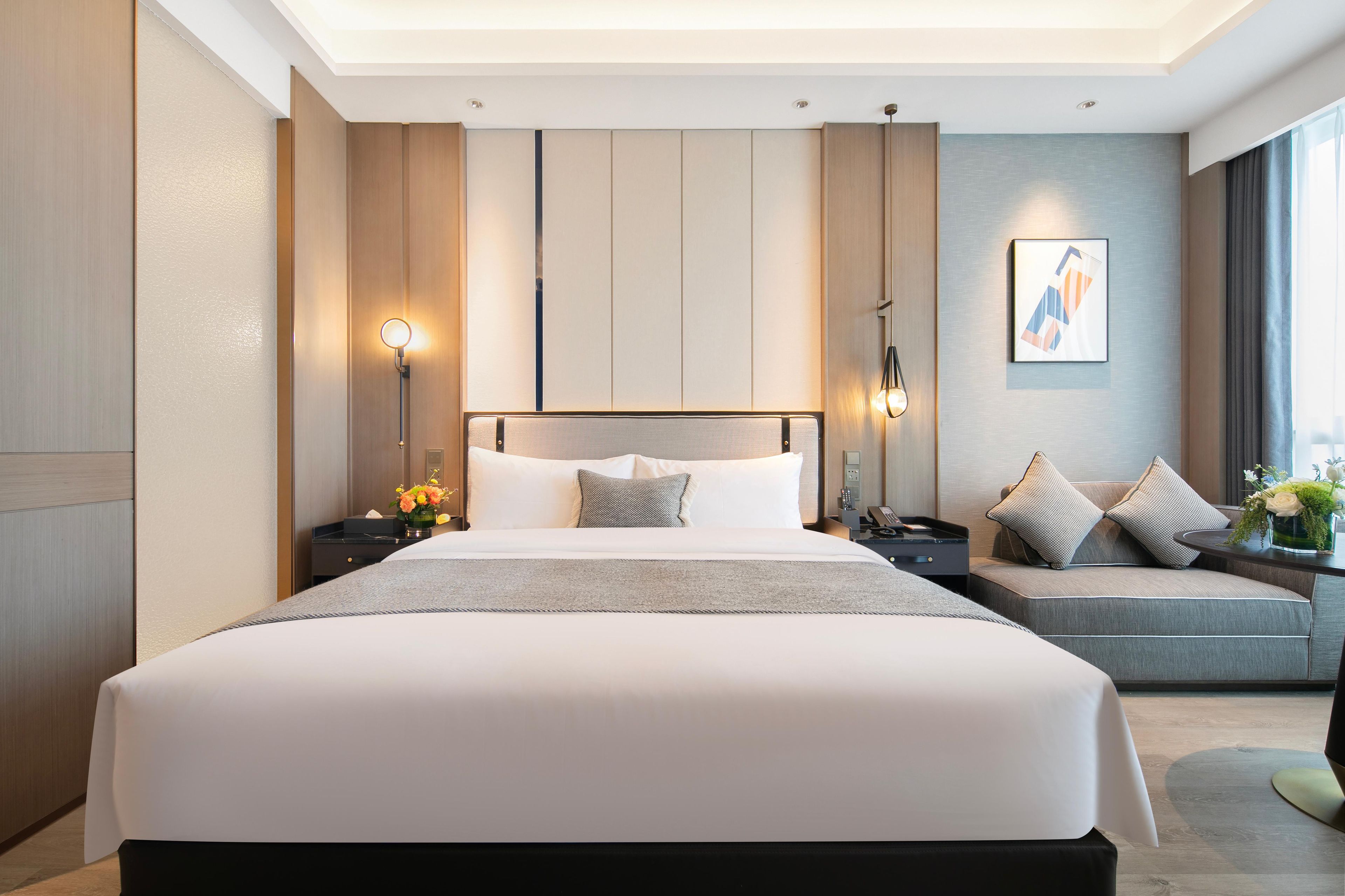 A modern hotel room with a large bed, bedside tables, a sofa, and a wall-mounted artwork.
