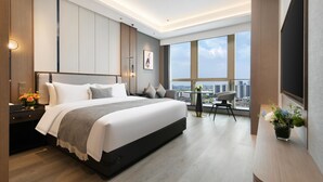 Een moderne hotelkamer met een groot bed, een bureau met stoel, een kleine tafel met stoel, en een groot raam met uitzicht op de stad.
