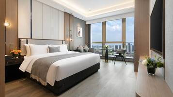 A modern hotel room with a large bed, a desk with a chair, a small table with a chair, and a large window offering a city view.