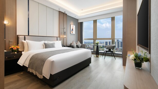 Een moderne hotelkamer met een groot bed, een bureau met stoel, een kleine tafel met stoel, en een groot raam met uitzicht op de stad.
