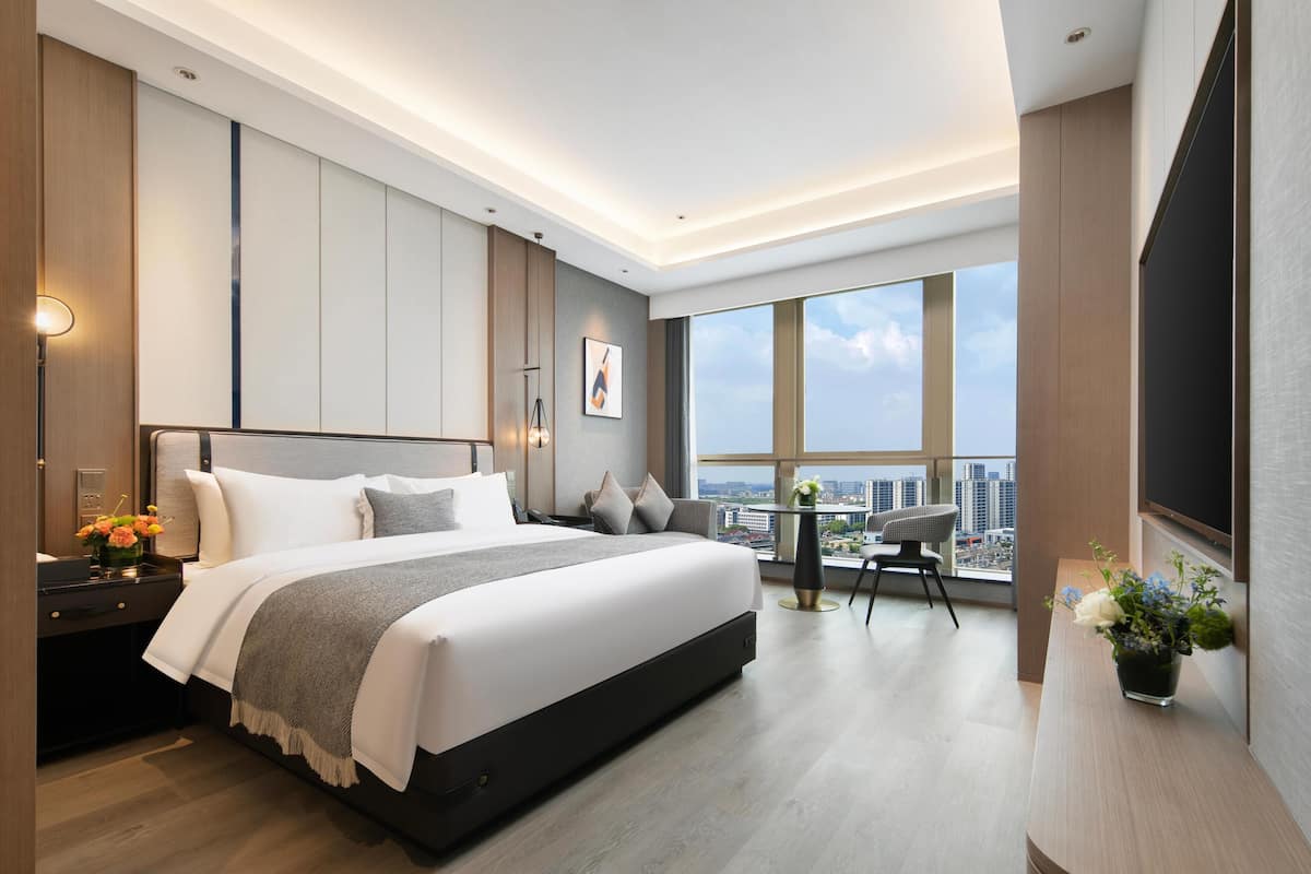 A modern hotel room with a large bed, a desk with a chair, a small table with a chair, and a large window offering a city view.