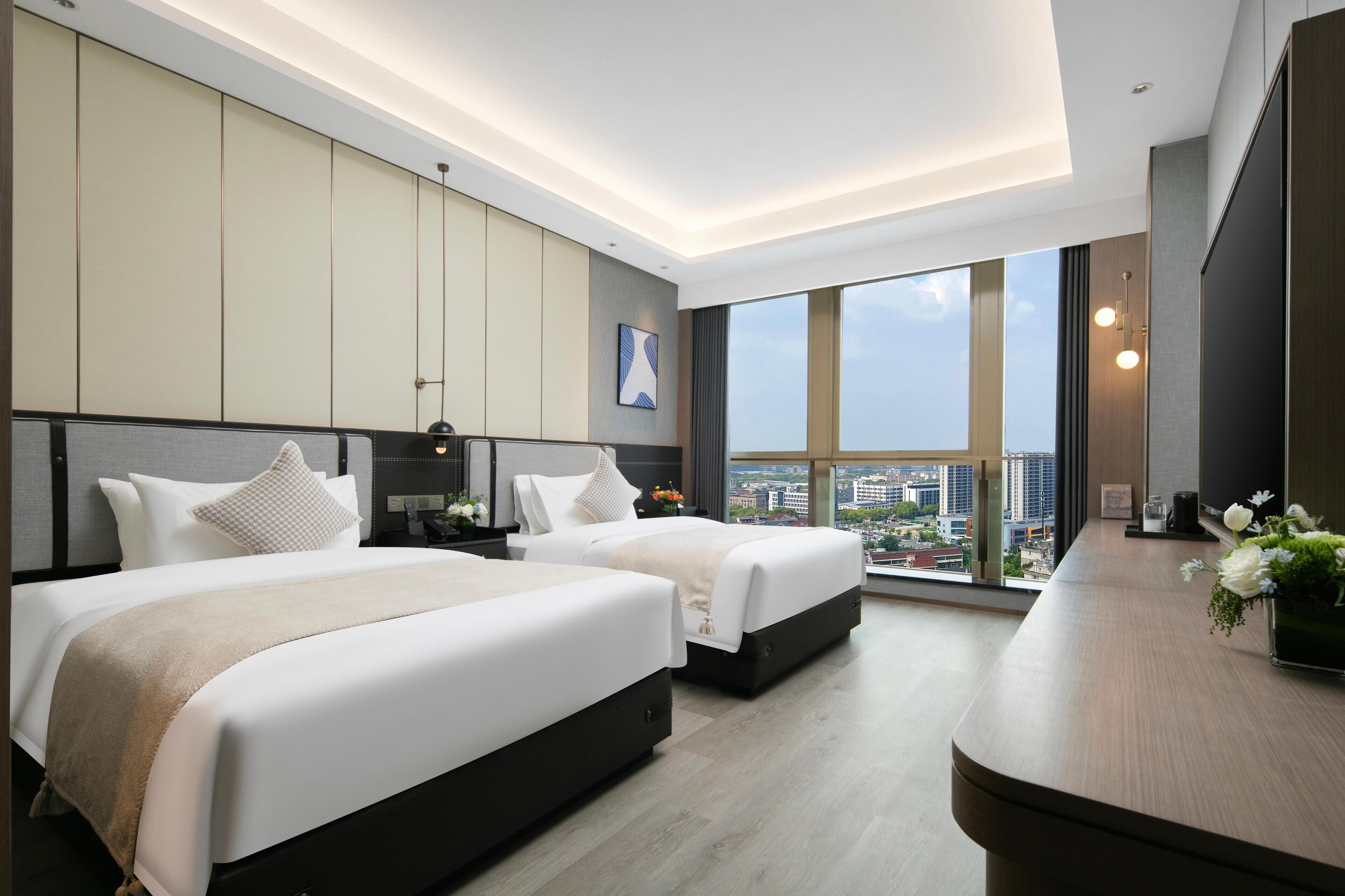 A modern hotel room with two beds, a large window with a city view, and a wooden desk with a vase of flowers.