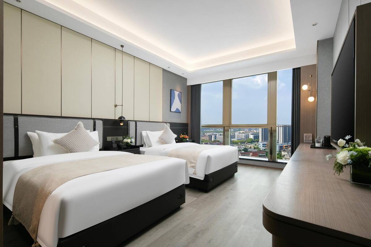A modern hotel room with two beds, a large window with a city view, and a wooden desk with a vase of flowers.