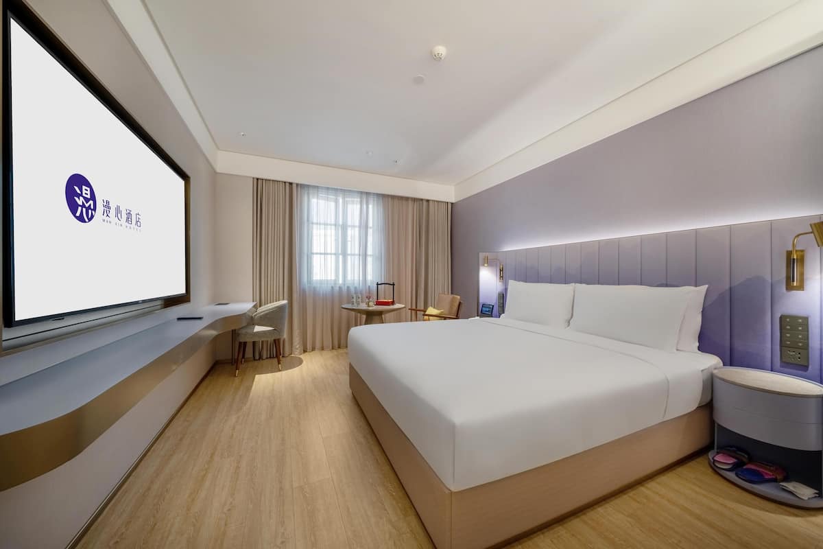 A modern hotel room with a large bed, a desk, and a TV screen displaying a logo.