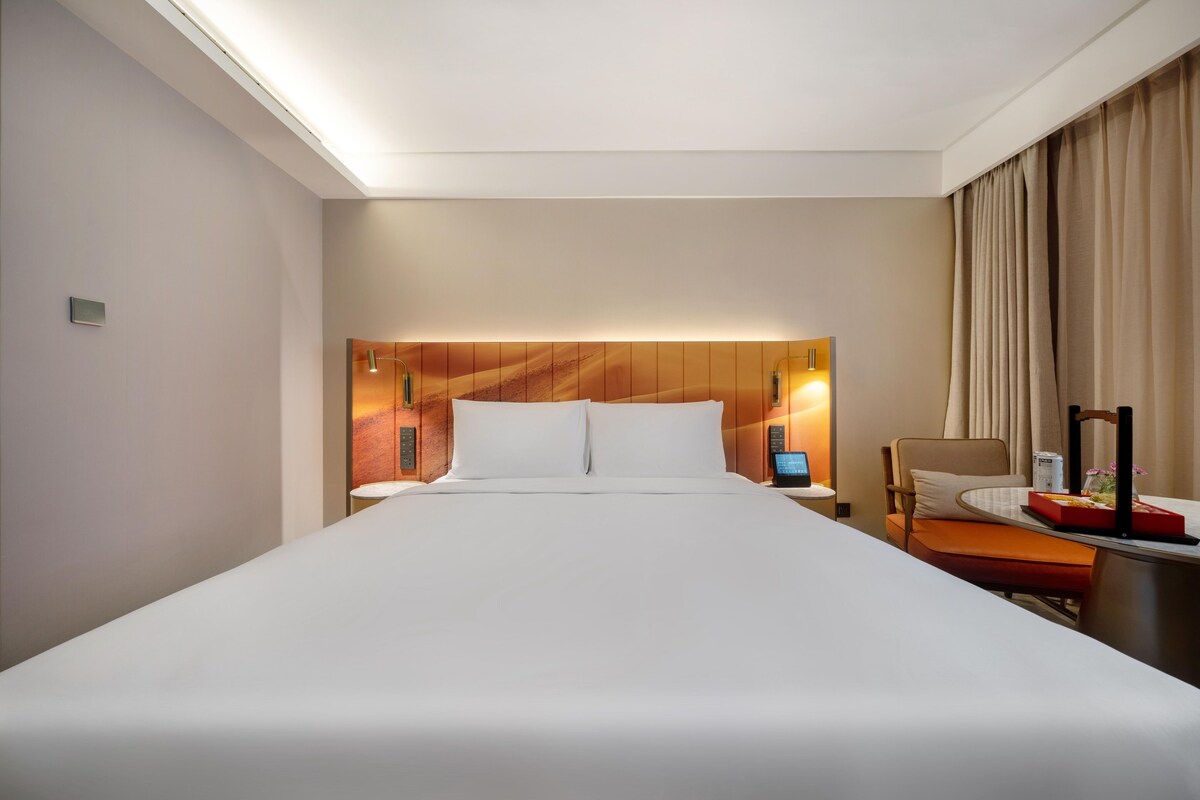 Habitación de hotel con una cama grande, un escritorio con computadora, una silla y una ventana con cortinas.