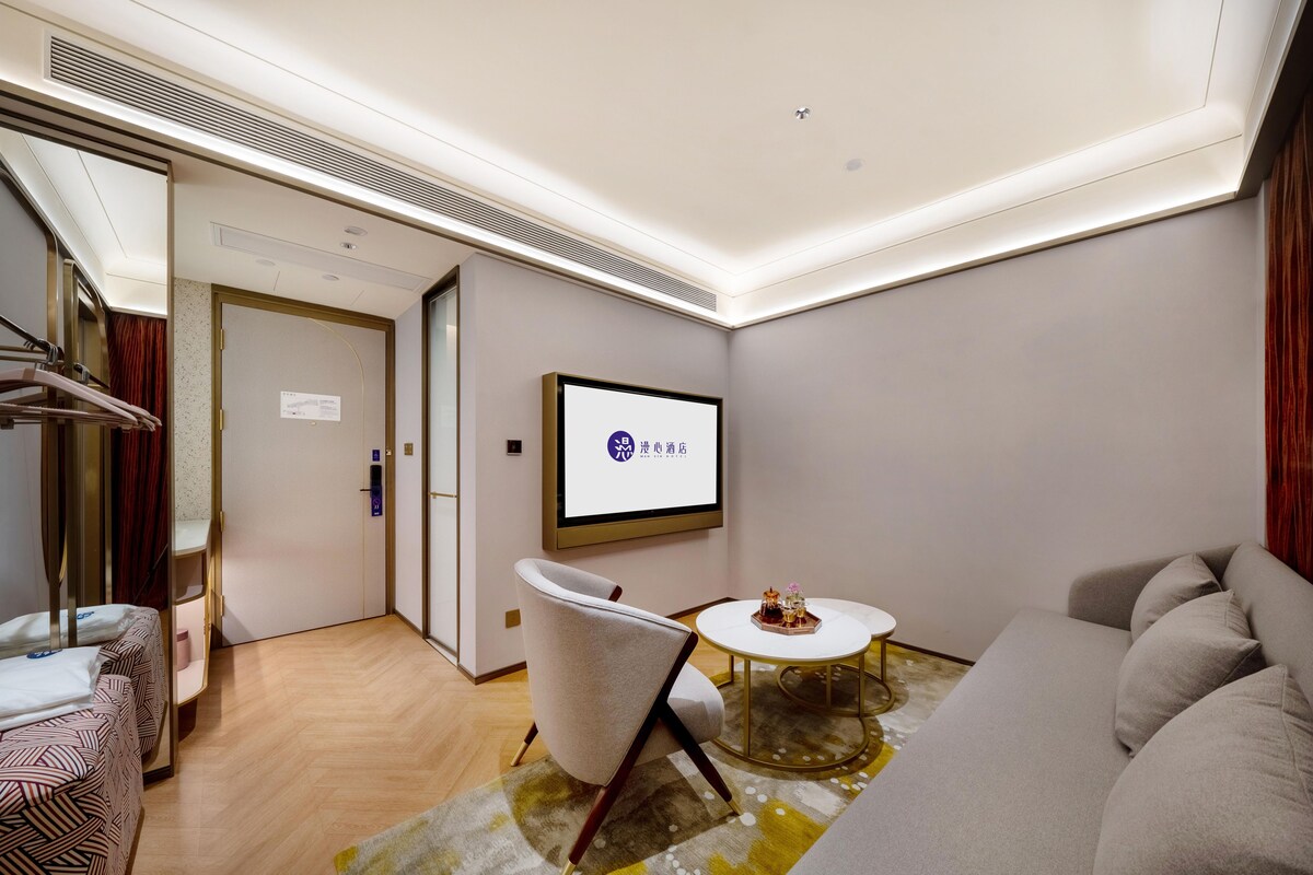 Una habitación de hotel moderna con un sofá, una silla, una mesita redonda pequeña, una cama con ropa de cama a rayas, un televisor montado en la pared y una puerta que da a otra habitación.