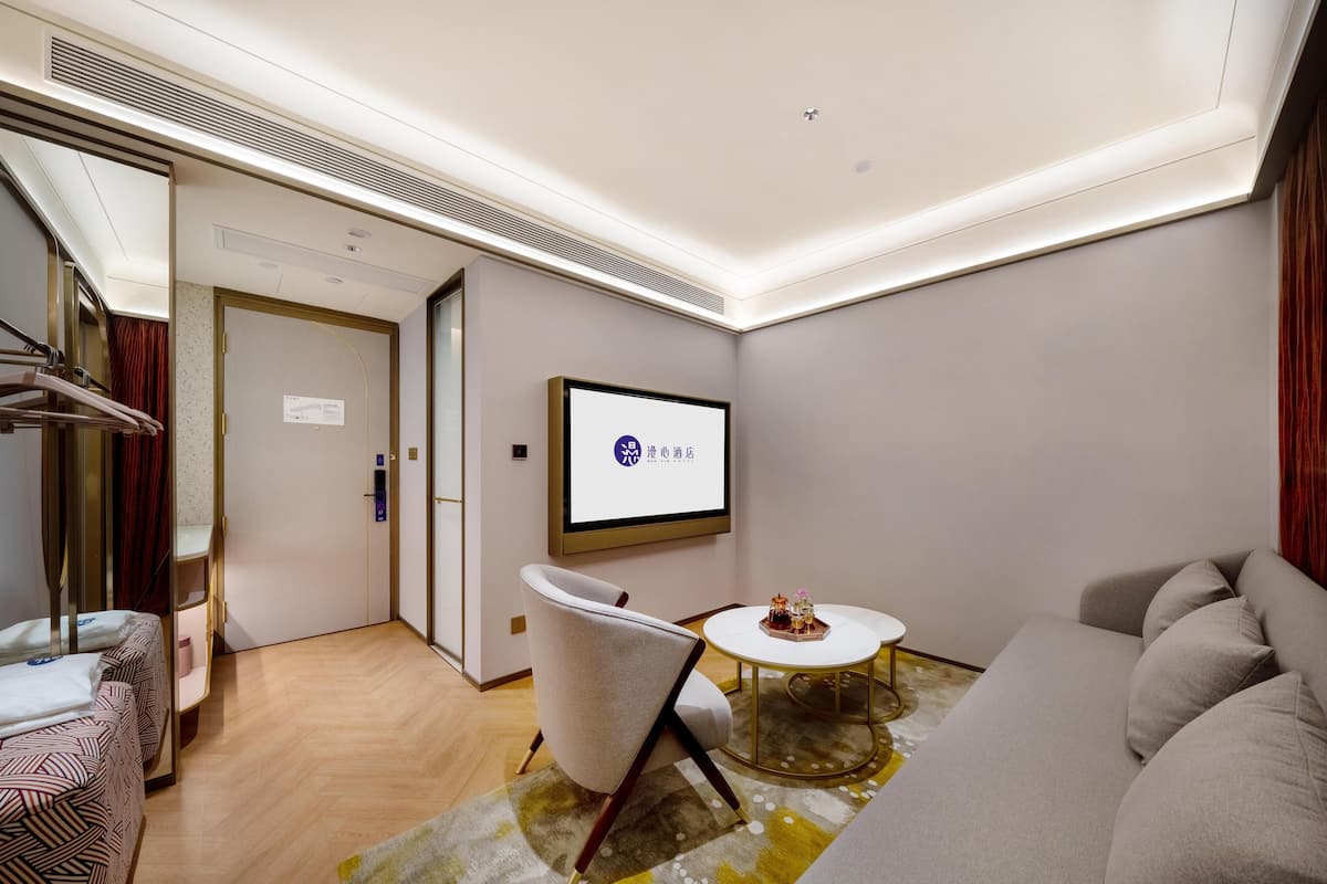 A modern hotel room with a sofa, a chair, a small round table, a bed with striped bedding, a wall-mounted TV, and a door leading to another room.