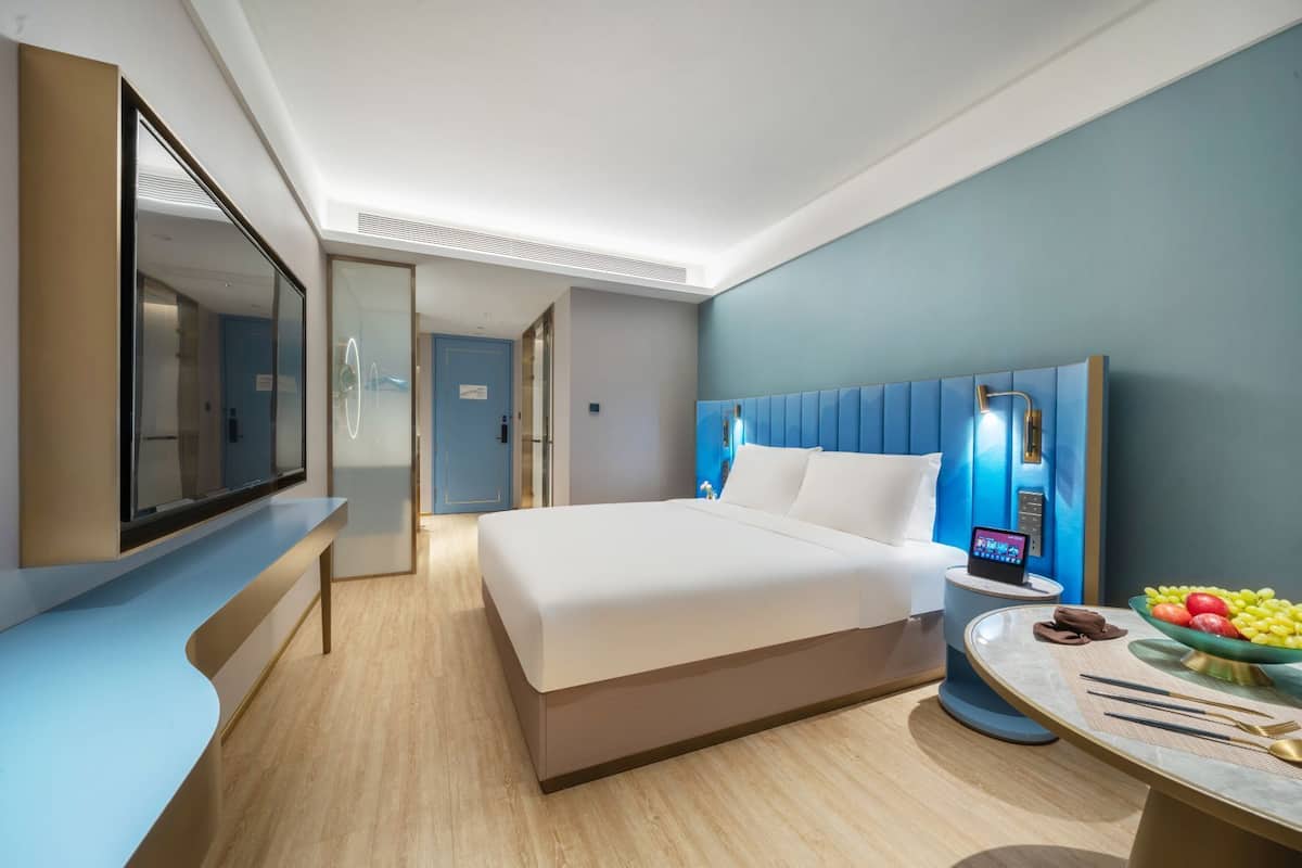 A modern hotel room with a large bed, a desk with a laptop, a bowl of fruit, and a mirror.