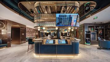 A modern lobby with a central reception desk, a digital information screen, and a staircase leading to an upper level.