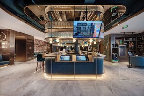 Un vestíbulo moderno con un mostrador de recepción central, una pantalla informativa digital y una escalera que conduce a un nivel superior.