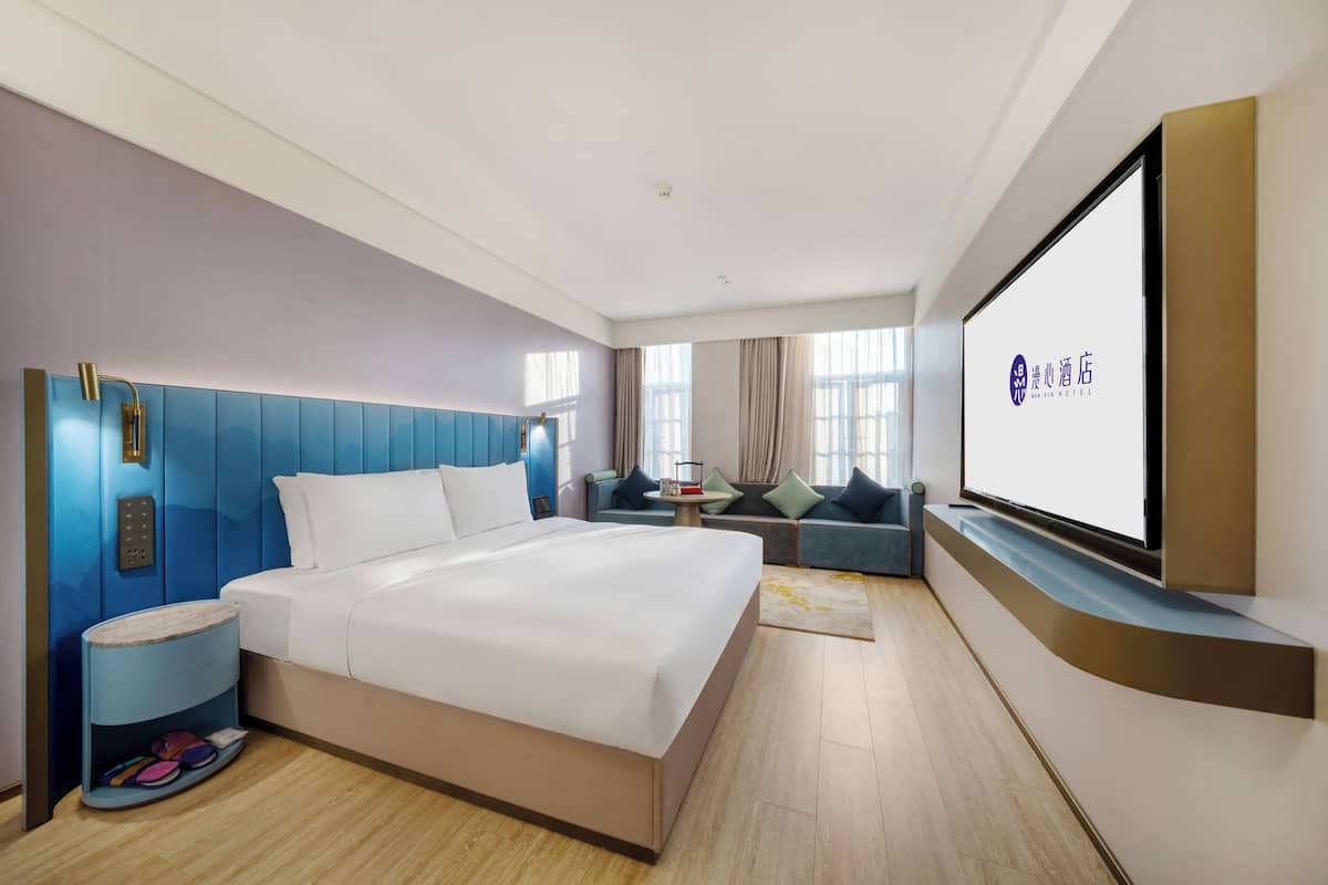 A hotel room with a bed, a TV, a seating area, and a wall-mounted screen displaying the hotel name.