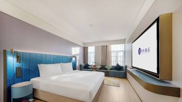 A hotel room with a bed, a TV, a seating area, and a wall-mounted screen displaying the hotel name.