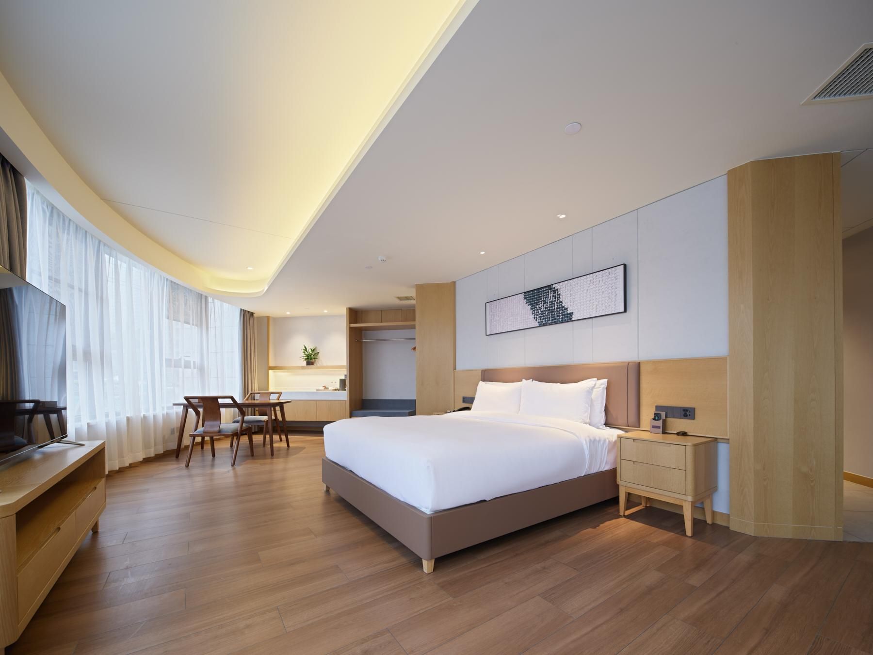 A modern hotel room with a large bed, a desk, and a chair. There is a small kitchenette in the corner.