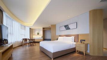 A modern hotel room with a large bed, a desk, and a chair. There is a small kitchenette in the corner.