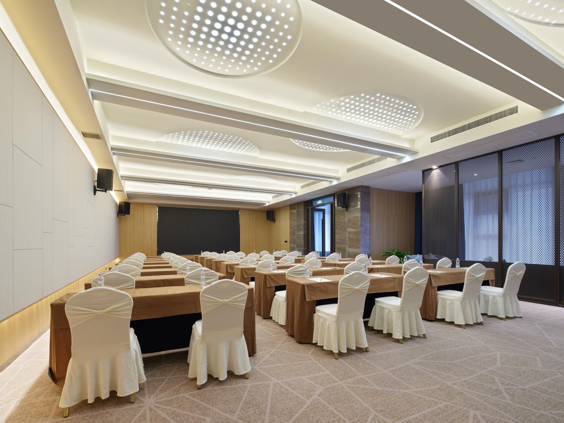 A conference room with rows of tables and chairs, a large screen, and a modern ceiling design.