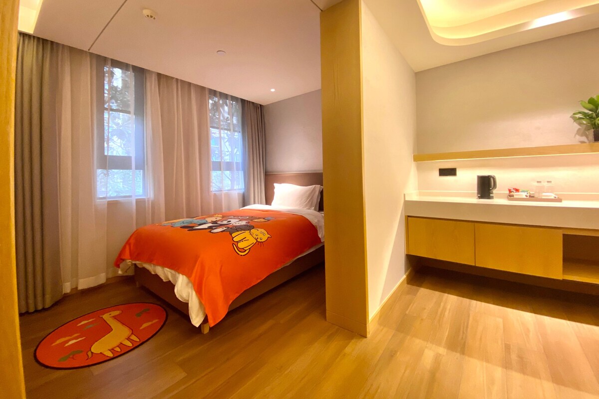 A hotel room with a bed, a bedside table, a window with curtains, a small plant on the table, and a wooden floor.