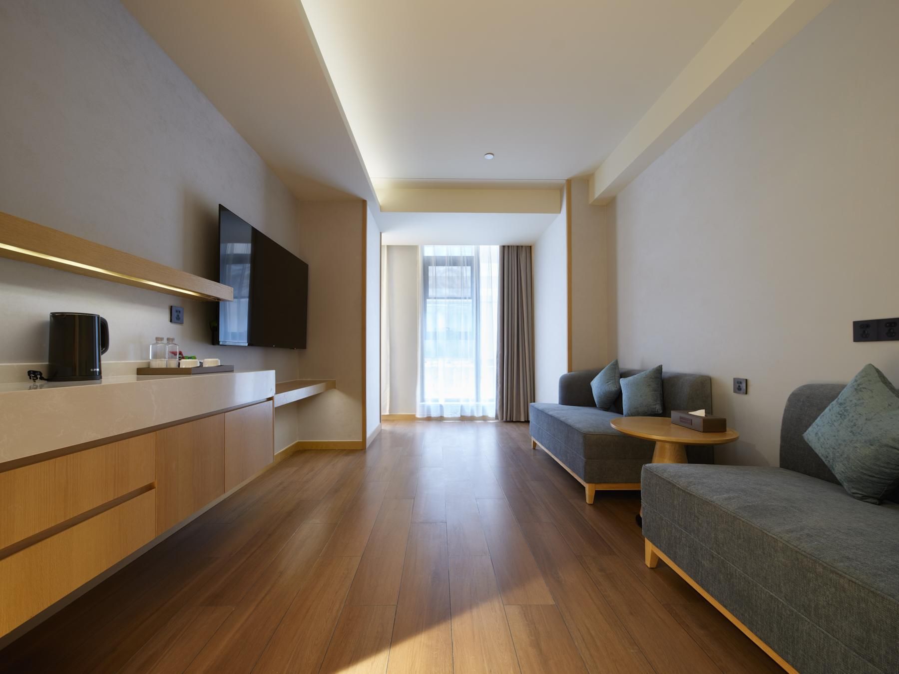 Una camera d'albergo moderna con pavimento in legno, un divano, un tavolino rotondo, una TV a parete e una finestra con tende.