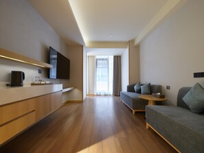 Une chambre d’hôtel moderne avec un plancher en bois, un canapé, une petite table ronde, un téléviseur fixé au mur et une fenêtre avec des rideaux.