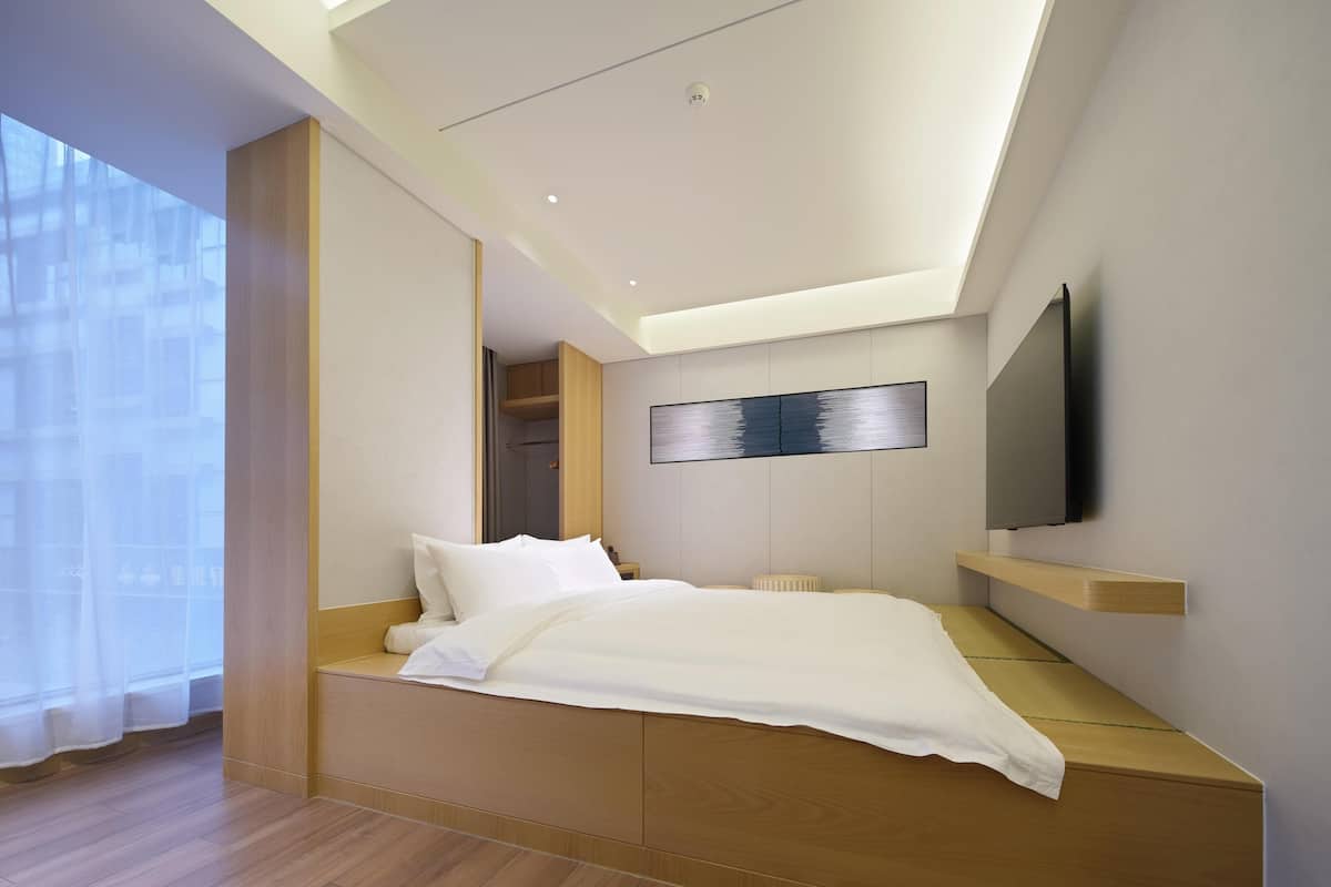 Ein modernes Schlafzimmer mit Bett, eingebauten Regalen, einem an der Wand montierten Fernseher und einem großen Fenster.