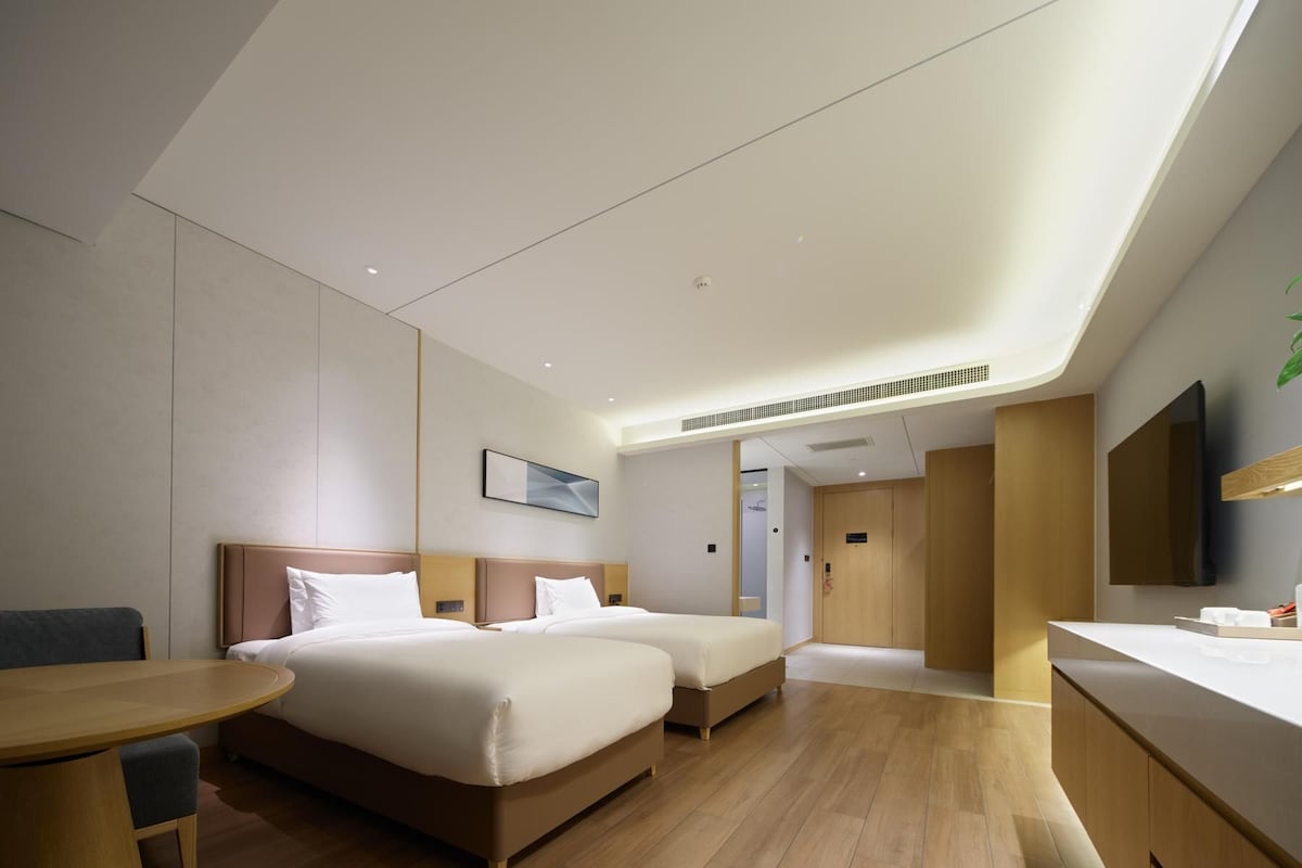 A modern hotel room with two beds, a desk, a chair, a TV, and a bathroom visible through an open door.
