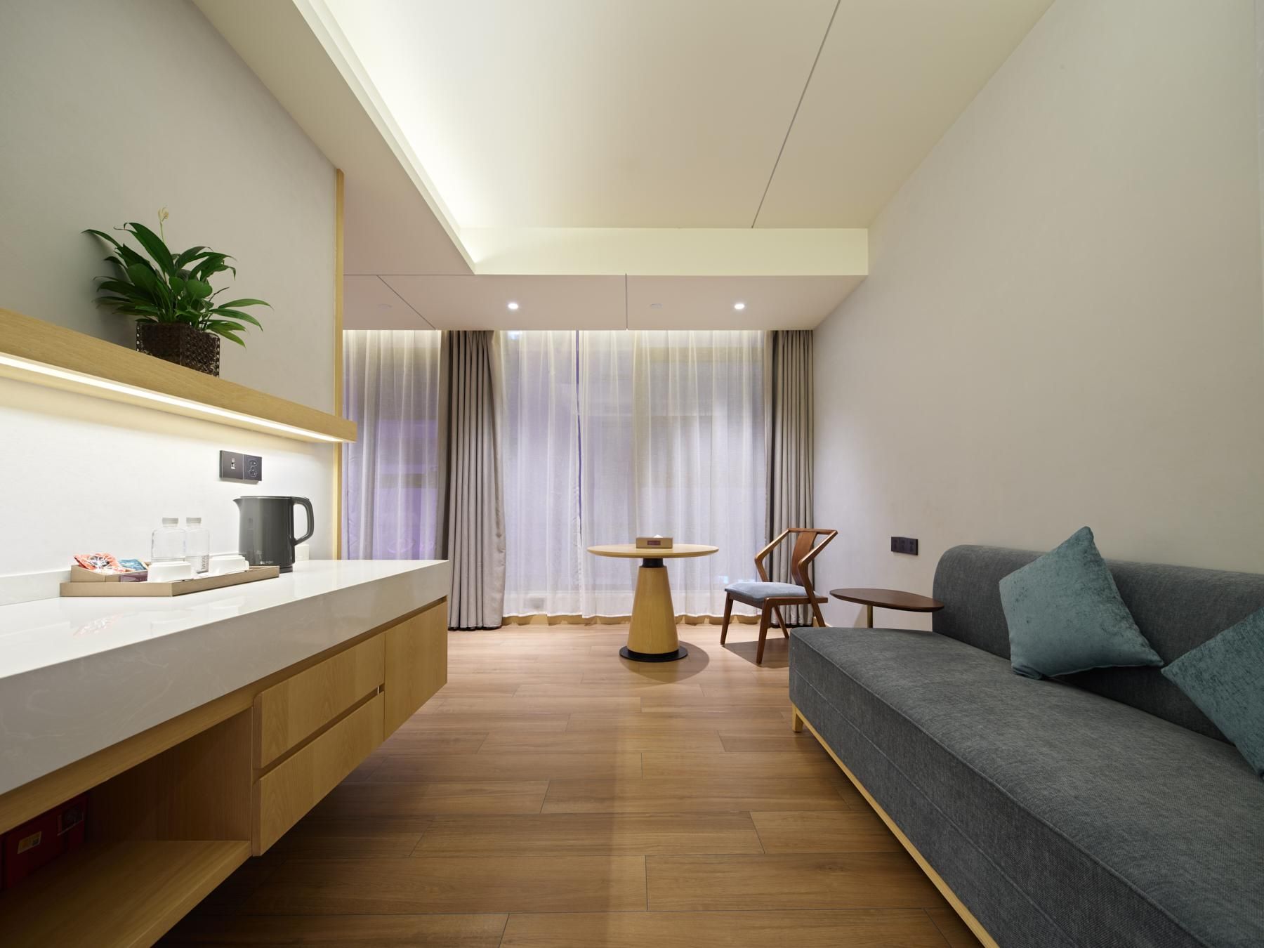 A modern hotel room with a wooden floor, a grey sofa, a small table, and a plant on the wall.