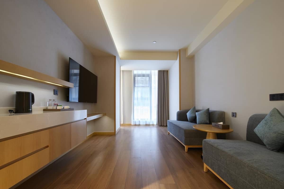 Ein modernes Hotelzimmer mit Holzboden, einer Couch, einem kleinen runden Tisch, einem an der Wand montierten Fernseher und einem Fenster mit Vorhängen.