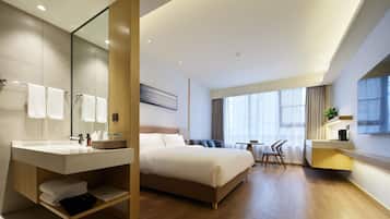 A modern hotel room with a large bed, a bathroom with a vanity, a dining area with a table and chairs, and a large window providing natural light.