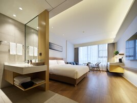 Habitación de hotel moderna con una cama grande, un baño con tocador, una zona de comedor con mesa y sillas, y un ventanal que proporciona luz natural.