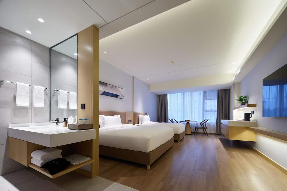 Ein modernes Hotelzimmer mit einem großen Bett, einem eigenen Badezimmer mit Waschtisch, einer Sitzecke mit Tisch und Stühlen sowie einem großen Fenster mit Aussicht.