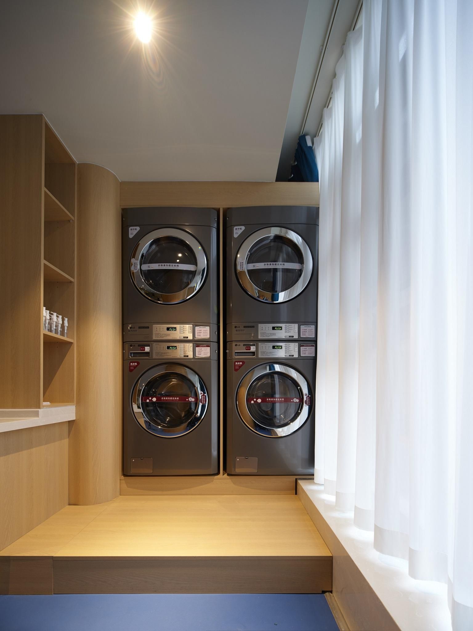 A compact laundry room with two wall-mounted washing machines, a wooden shelf with toiletries, and a window with white curtains.