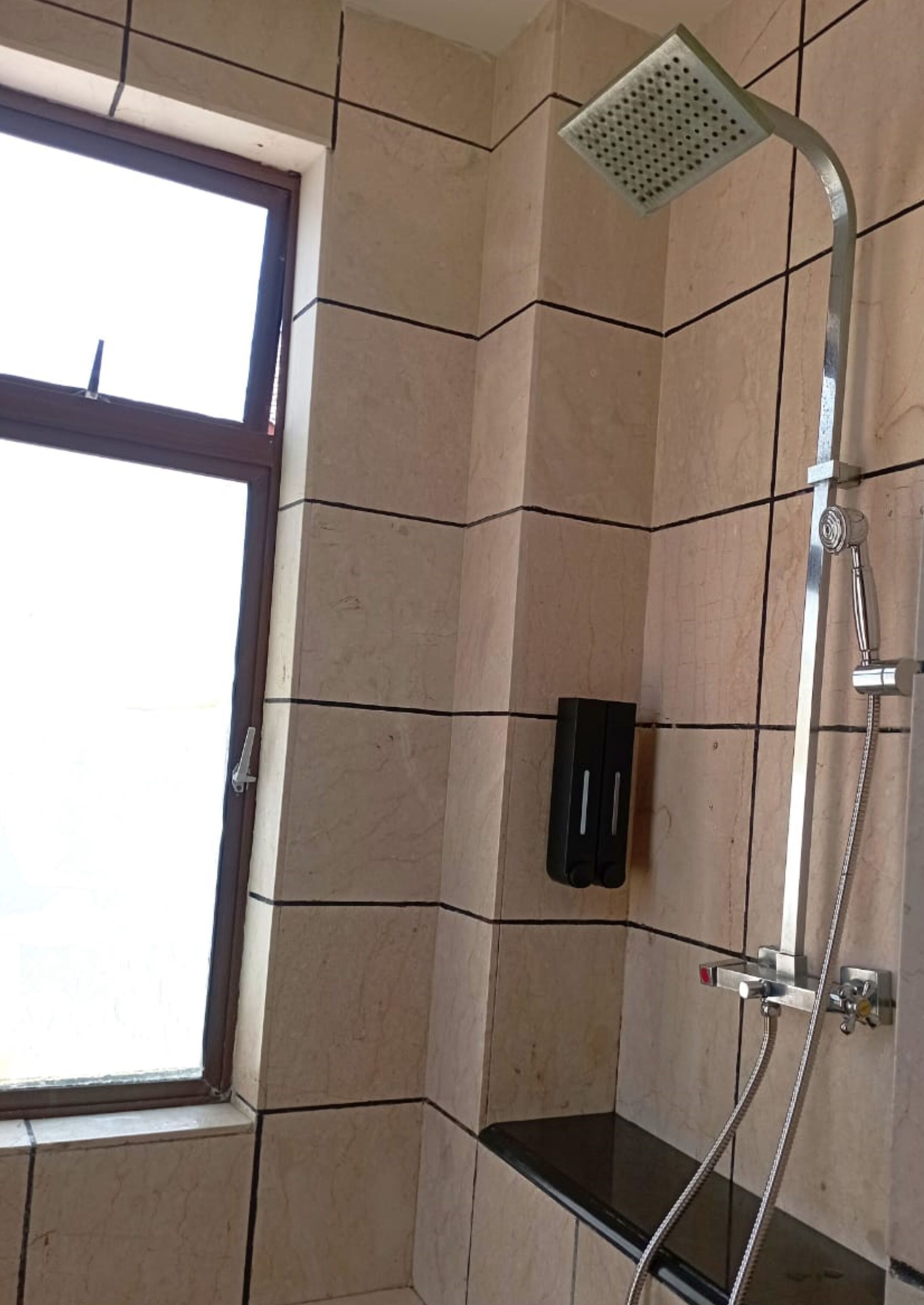 Zona de ducha con cabezal de ducha de mano, lavamanos con cerramiento de vidrio y una ventana.