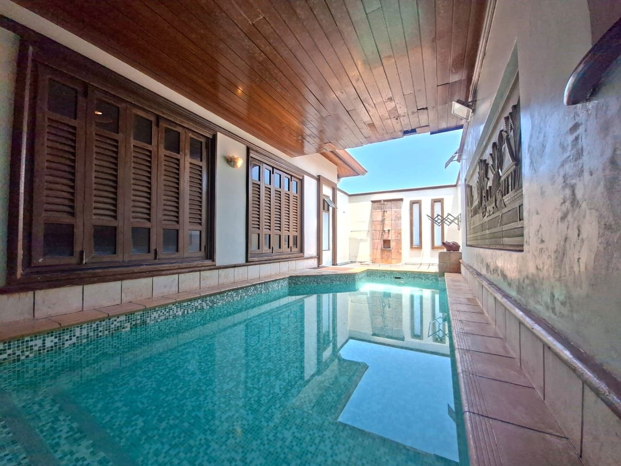 Una piscina con bordo piastrellato, soffitto in legno e vista su un edificio dalla facciata piastrellata e una porta.