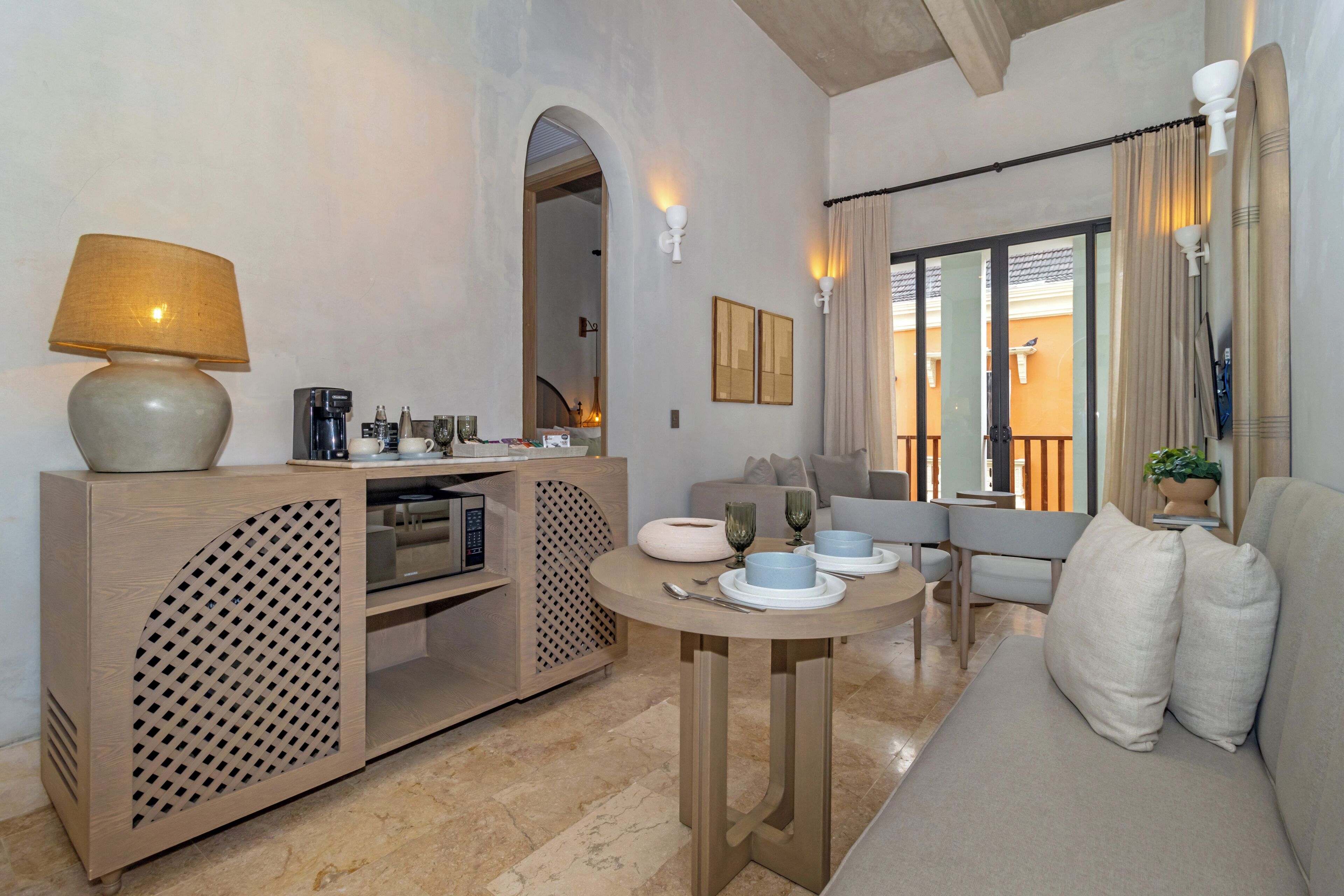 A modern living room with a sofa, a round table, a coffee station, and a view of the outside.