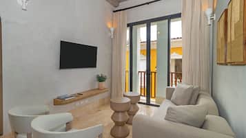 A modern living room with a flat-screen TV, a sofa, two chairs, a small table with a plant, and a sliding glass door leading to an outdoor balcony.