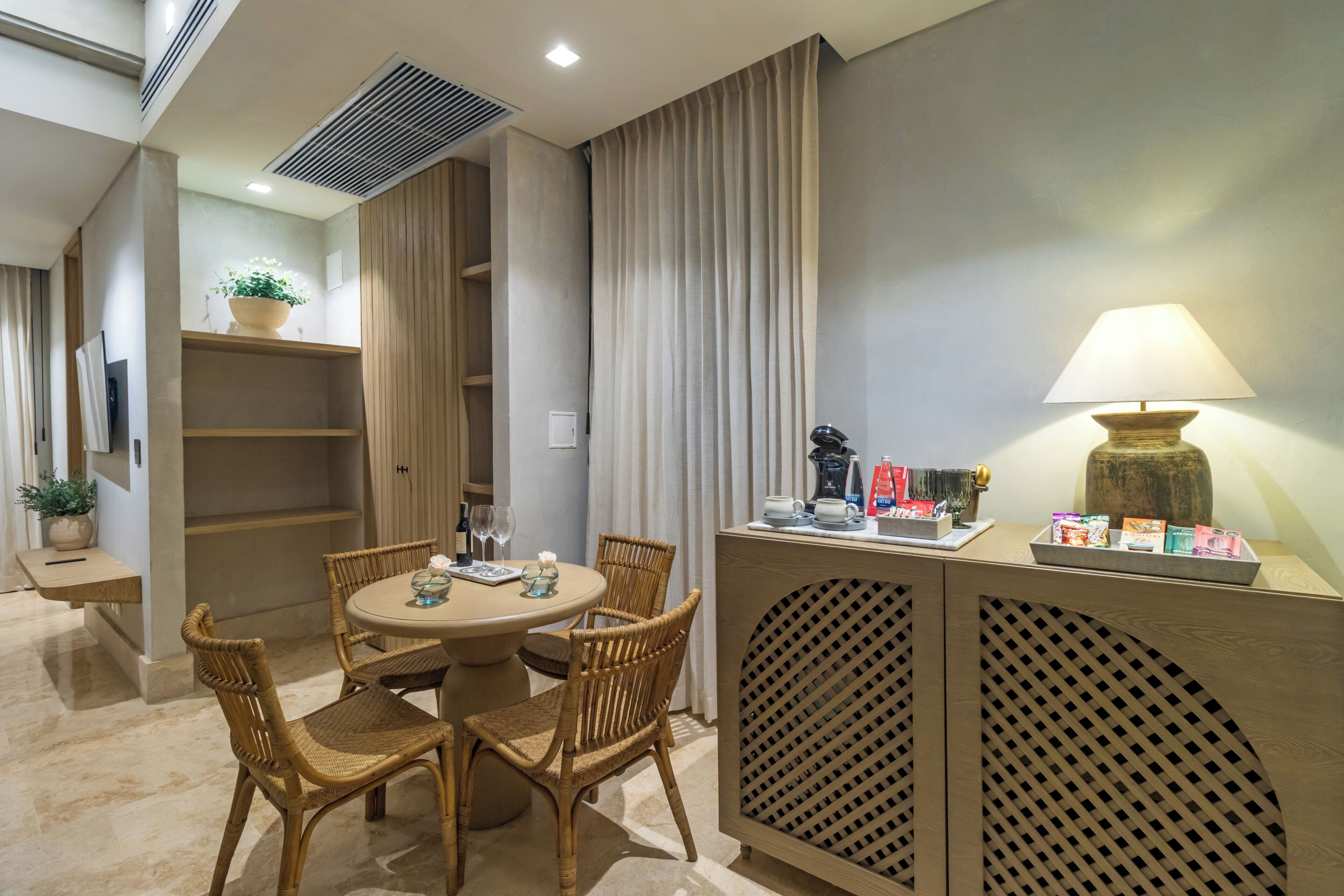 A dining area with a round table, wicker chairs, a cabinet with a lamp, and a potted plant.