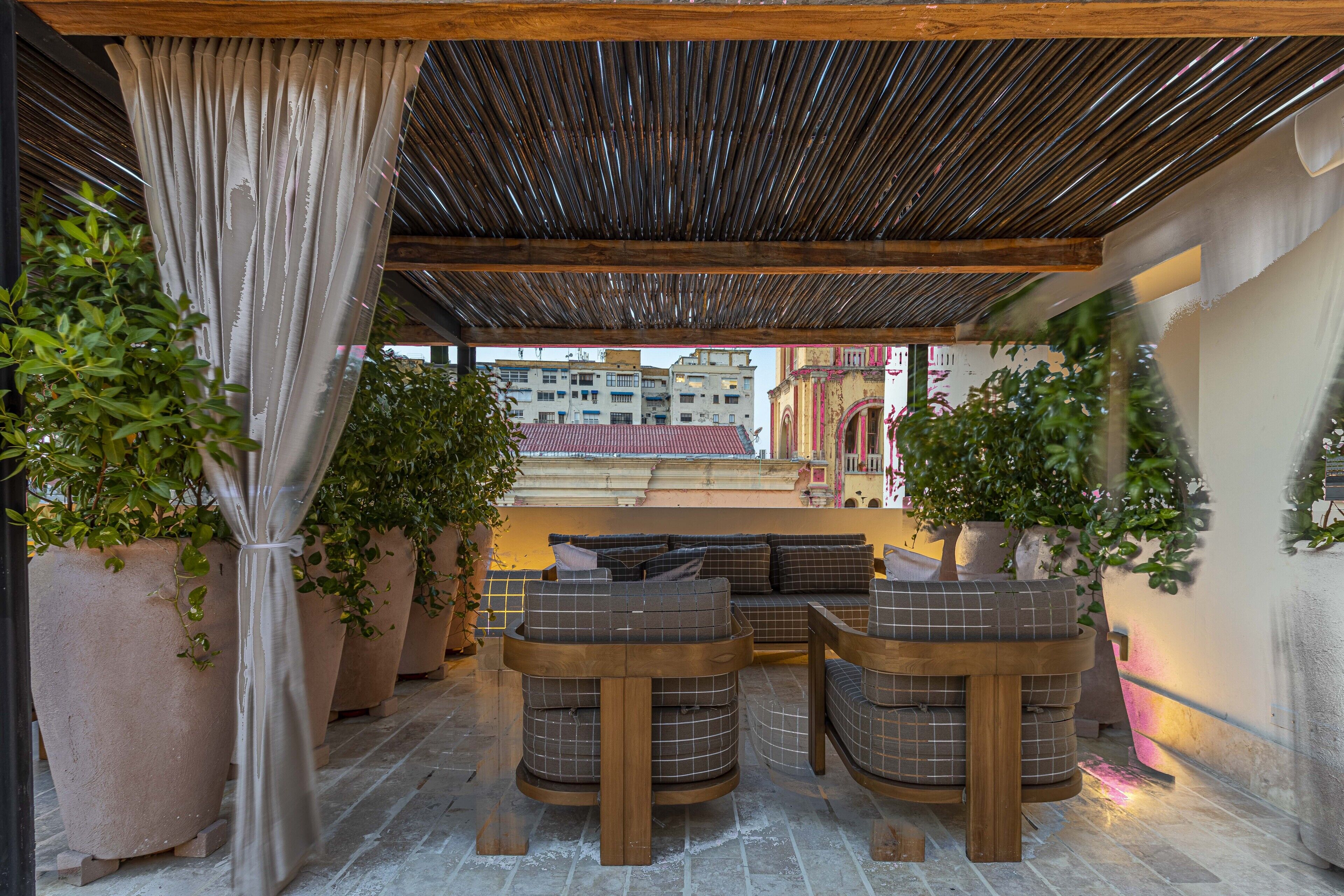 An outdoor seating area with a wooden bench and a cushioned sofa, surrounded by large potted plants.