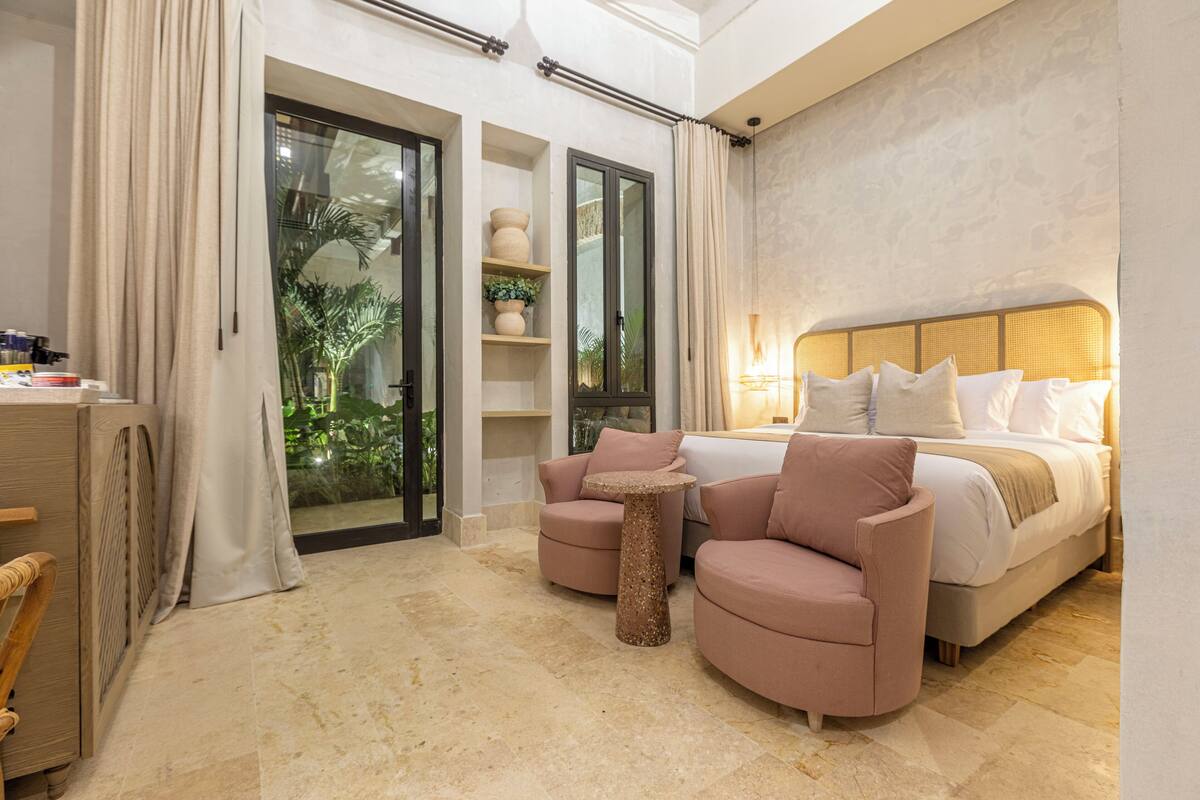 A hotel room with a bed, two armchairs, a small table, a desk, a plant, and a view through a glass door.