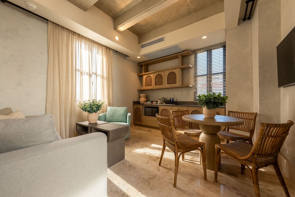A modern living room with a sofa, a dining table with chairs, a kitchen area with cabinets, and a large window with curtains.