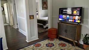 A hotel room with a large flat-screen TV mounted on the wall, a wooden cabinet below it, a patterned rug on the floor, and a bed with a green bedspread in the background.