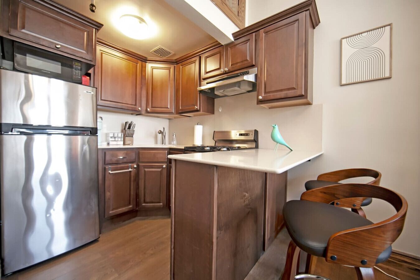 Una cocina con armarios de madera, electrodomésticos de acero inoxidable, una encimera blanca y dos sillas de madera.