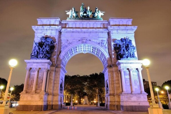 A large, illuminated archway with statues on top, surrounded by trees and street lamps.