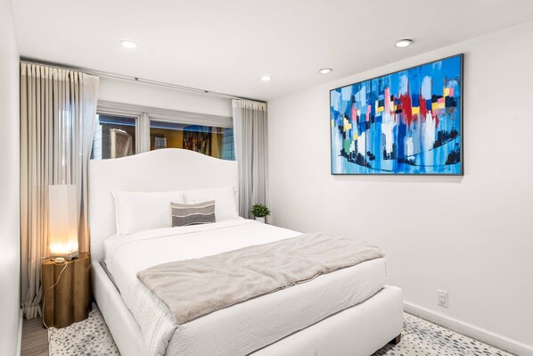 Un dormitorio moderno con una cama grande, una mesita de noche, un cuadro colgado en la pared y una ventana con cortinas.