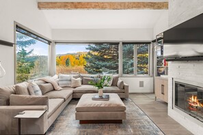 Una sala moderna con un amplio sofá modular, una chimenea y vistas del follaje otoñal a través de amplios ventanales.