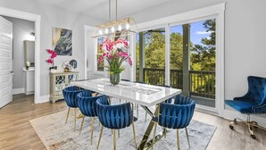 A dining area with a white table, blue velvet chairs, a chandelier, and a view of trees through large windows.