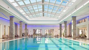 A large indoor swimming pool with a glass ceiling, surrounded by lounge chairs and a view of palm trees.