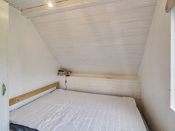 A small, white, sloped-roofed room with a single bed, a shelf with books and a small lamp, and a window on the right.