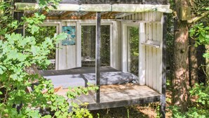 Een houten hangstoel met metalen frame, omgeven door bomen en begroeiing.