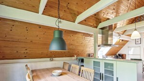 Een houten eettafel met stoelen, een hanglamp en een keuken met kasten en apparatuur.