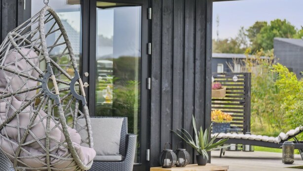 Un espacio exterior moderno con un sillón colgante de mimbre, una silla acolchada y una mesa de madera con plantas en macetas y faroles.