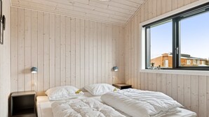 Un dormitorio con pared de paneles de madera, un ventanal grande, una cama con ropa de cama blanca, dos mesitas de noche con lámparas y un estante pequeño.